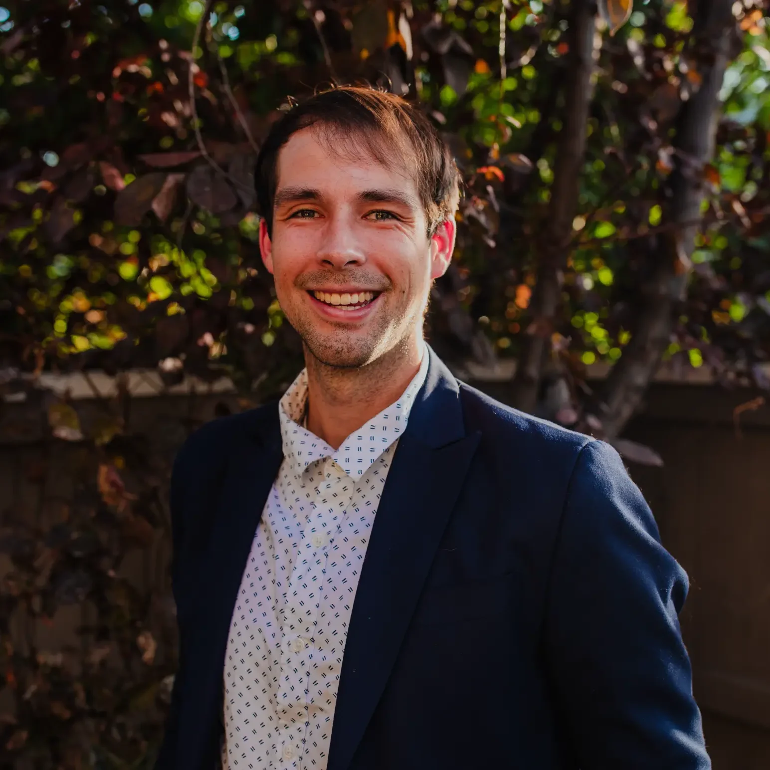 Professional outdoor portrait of counselor Hank Shuler smiling and wearing a navy blazer and patterned button-up shirt with warm natural light and blurred trees in the background.